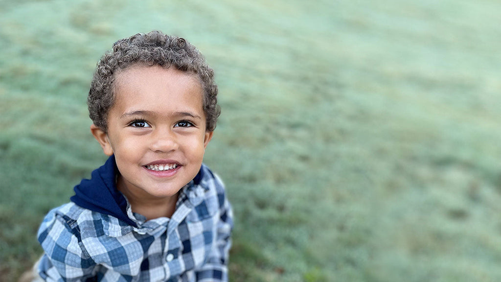 Brown curly haired boy looking up at the camera and smiling while wearing a blue plaid shirt with a blue hood while sitting on a green grass field