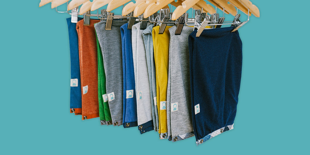 Close up of 10 hoods in a variety of colors with Thoughtfully Hooded patch hanging from pant hangers on a teal background