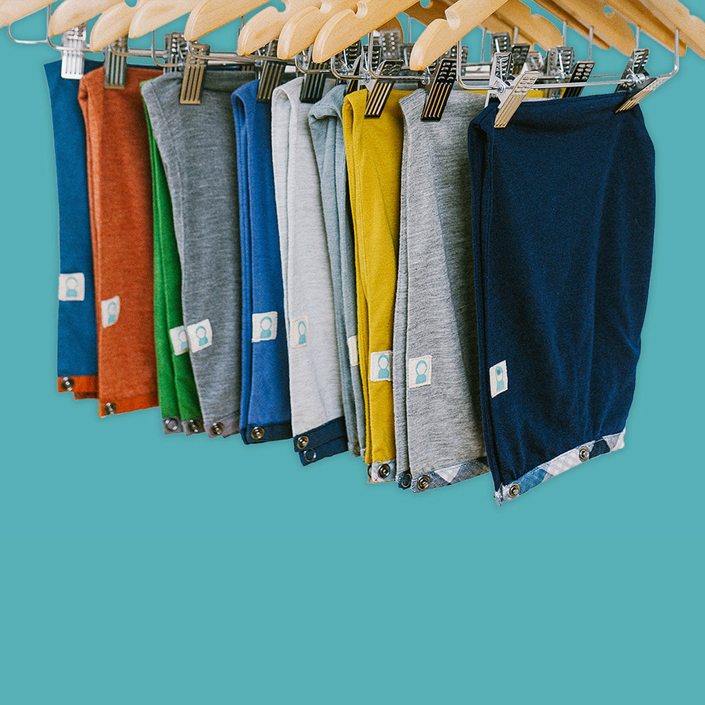 Close up of 10 hoods in a variety of colors with Thoughtfully Hooded patch hanging from pant hangers on a teal background