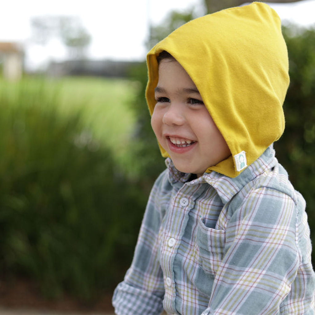 child wearing yellow hood and button up plaid shirt. Background is out of focus, green field with bushes